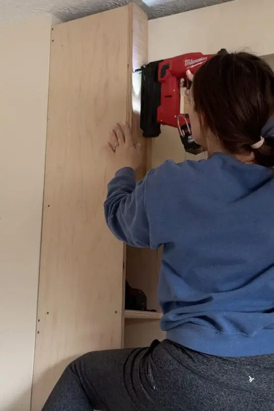 A woman in a blue sweatshirt using a red Milwaukee cordless nail gun to attach a side panel to a built-in shelving unit. She is kneeling on a step stool, ensuring the panel is flush against the structure for a secure fit.